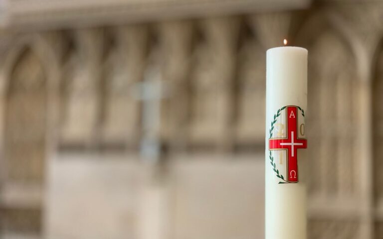 Burning white candle with red cross pattern on blurred background of church stone wall